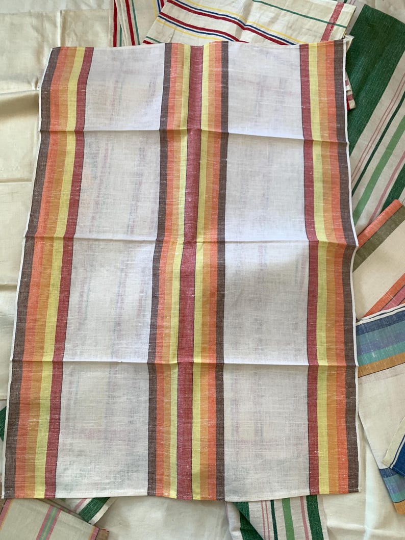 May include: White linen towel with vertical stripes in shades of orange, yellow, and brown. Other striped towels in various colors are visible in the background. The towel is folded and appears to be new.