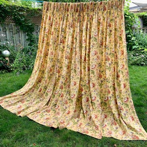 May include: A floor-length curtain featuring a yellow background with a dense floral pattern in shades of red, pink, green, and blue. The fabric is gathered at the top with pleats, creating vertical folds that drape down to the ground.