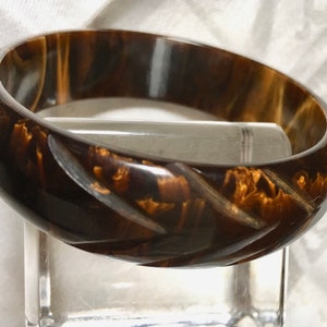 May include: A vintage brown bangle bracelet with a carved diagonal pattern. The bracelet has a glossy finish and a marbled appearance with shades of brown and gold. This accessory is a classic piece of jewelry.