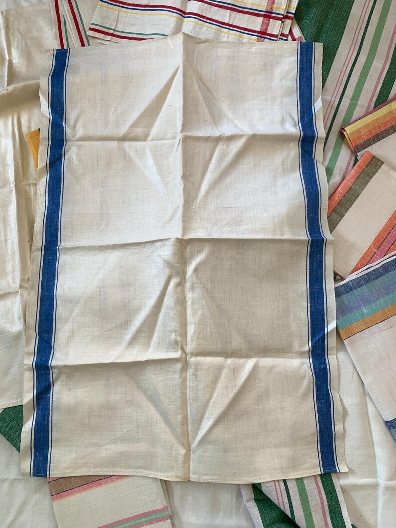 May include: Cream-colored linen kitchen towel with a blue stripe border. Several other striped towels in various colors are visible in the background. The main towel is folded and has visible creases.