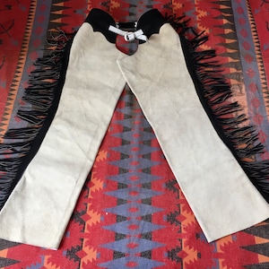 May include: A pair of beige suede chaps with black fringe along the sides. The chaps have a black waistband and a silver buckle closure. The chaps are laid out on a patterned red, blue, and brown rug.