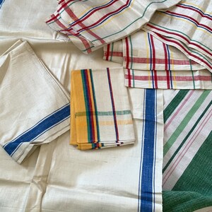 May include: Assortment of vintage linen textiles in various patterns. Includes white fabric with blue stripes, folded napkins with colorful stripes, and a green and pink striped fabric. The textiles are arranged on a light-colored surface.