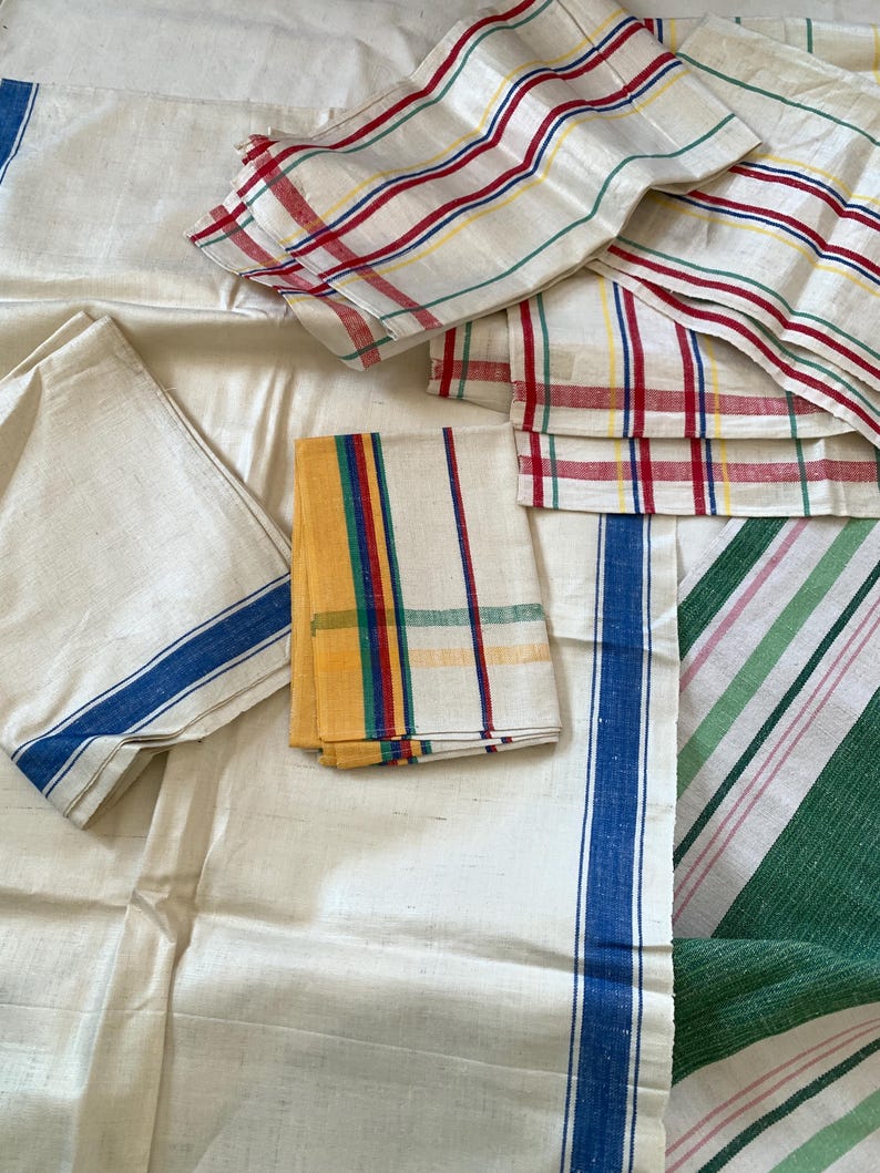 May include: Assortment of vintage linen textiles in various patterns. Includes white fabric with blue stripes, folded napkins with colorful stripes, and a green and pink striped fabric. The textiles are arranged on a light-colored surface.