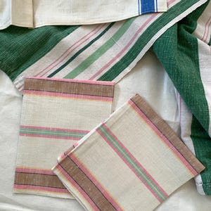 May include: A collection of linen kitchen towels and napkins. The towels feature stripes in green, pink, and blue. Napkins have pink, brown, and yellow stripes. The textiles are arranged on a white surface.