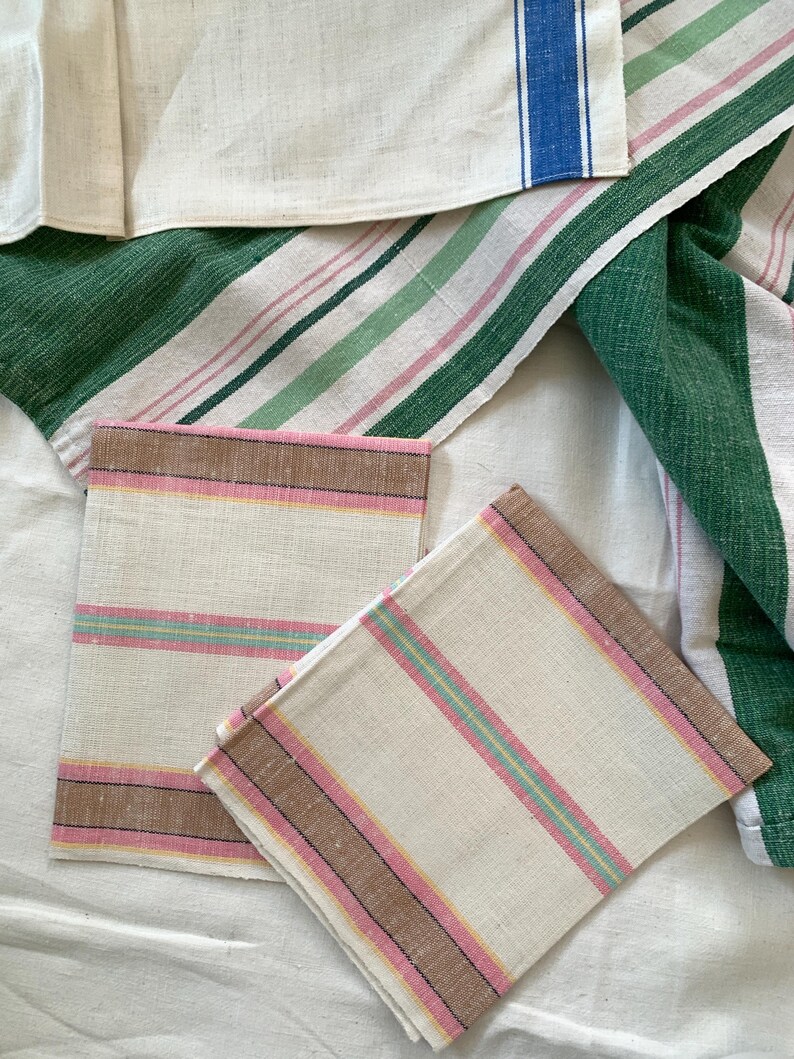 May include: A collection of linen kitchen towels and napkins. The towels feature stripes in green, pink, and blue. Napkins have pink, brown, and yellow stripes. The textiles are arranged on a white surface.