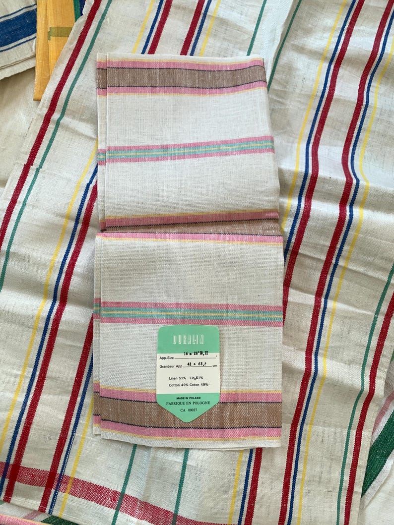 May include: White linen and cotton blend hand towel with pink, brown, and green stripes. The towel has a tag that reads "App. Size 16" x 16", Grandeur App. 41 x 41 cm, Linen 51%, Cotton 49%" and "Made in Poland, Fabrique en Pologne, CA 00027."