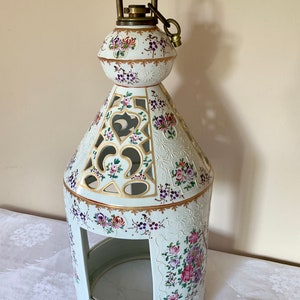 May include: A white ceramic lantern with floral designs in pink, purple, and green. The lantern has a decorative, openwork design with gold accents and a brass handle. The overall shape is a tall, ornate cylinder.
