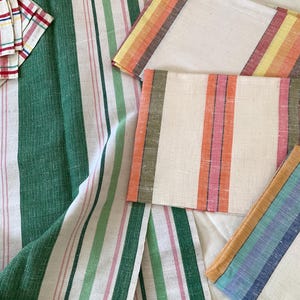 May include: Assortment of striped fabric items in various colors. Includes green, pink, and white striped fabric, along with several rectangular pieces featuring stripes in orange, yellow, and blue. The textiles appear to be linen or a similar natural fiber.
