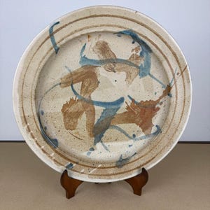 Może przedstawiać: Okrągły talerz ceramiczny o nakrapianej, kremowej podstawie, ozdobiony abstrakcyjnymi pociągnięciami pędzla w odcieniach brązu i błękitu. Talerz posiada szeroki, uniesiony brzeg z dwiema koncentrycznymi liniami oraz gładkie, szkliwione wykończenie.