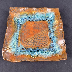 May include: A square ceramic plate with a textured, burnt orange center featuring a diagonal chevron pattern. The plate has a wide, uneven border with a reactive glaze in shades of bright turquoise and deep blue, creating a crystalline effect.