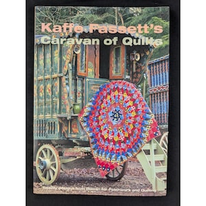 Puede incluir: Un libro de tapa blanda titulado Kaffe Fassett's Caravan of Quilts. La portada muestra una colcha geométrica de colores vibrantes sobre los escalones de una caravana de madera pintada y ornamentada. El texto está en letras blancas y rosas.