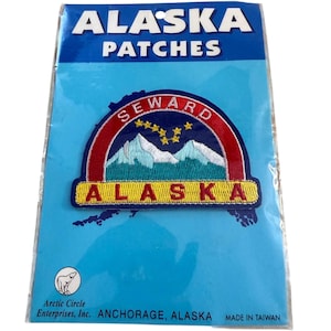 May include: An embroidered patch in a clear plastic package labeled ALASKA PATCHES. The patch features a red arch with the word SEWARD, a blue sky with yellow stars, white mountains, and a yellow base with the red text ALASKA.