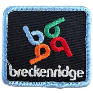 May include: A square embroidered patch with a black background and a light blue stitched border. The design features a stylized logo with light blue, green, and orange letters, and the word breckenridge stitched in white lowercase letters below.