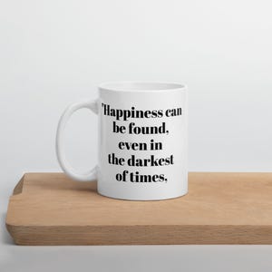 May include: A white ceramic mug featuring the black printed text, "Happiness can be found, even in the darkest of times,". The mug has a smooth, glossy finish, a curved handle on the left side, and a cylindrical body with a flat base for stability.
