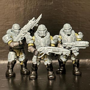 May include: Three gray plastic soldier action figures wearing full-body tactical armor and helmets. Each figure holds a detailed futuristic rifle, with the left figure's weapon featuring an attached bayonet. The figures have dark boots.