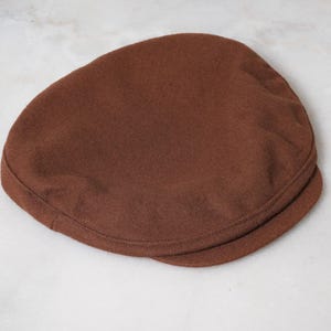 May include: A solid brown flat cap featuring a rounded crown and a short, stiff brim. The hat is constructed from a soft, felted fabric with visible stitching along the edge of the brim and the base of the crown, creating a structured shape.
