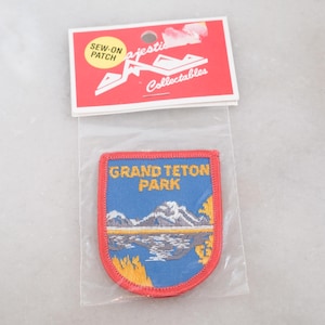 May include: A blue sew-on patch featuring an embroidered landscape of Grand Teton Park with mountains, water, and yellow foliage. The patch is enclosed in a clear plastic bag with a red header card labeled Majestic Collectables and SEW-ON PATCH.
