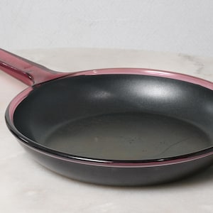 May include: A round frying pan with a matte black interior cooking surface and a translucent deep magenta glass rim and handle. The pan features a smooth, circular design with a long, integrated handle extending from the side of the vessel.