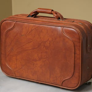 May include: A rectangular brown suitcase with a smooth, marbled texture and a curved, stitched border design. The luggage features a sturdy top handle with a knotted center detail and a structured frame with visible metal rivets on the side panel.