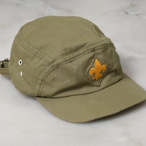 May include: An olive green five-panel cap featuring a yellow embroidered fleur-de-lis emblem on a diamond-shaped patch at the front. The hat includes two white metal ventilation eyelets on the side and an adjustable strap with a metal buckle.