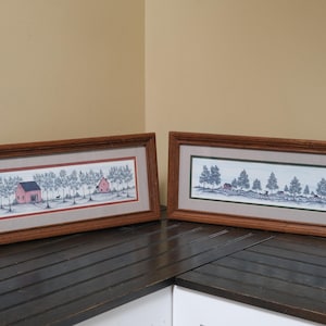 May include: Two rectangular framed landscape prints featuring ink-style drawings of rural scenes with trees and small buildings. The left print displays red buildings, while the right print shows a red building and a horse-drawn carriage in a snowy setting.