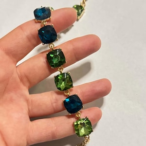 Könnte beinhalten: Ein goldfarbenes Kettenarmband mit abwechselnd quadratisch geschliffenen Kristallen in Dunkelblau und leuchtendem Grün. Das Armband besteht aus einer goldfarbenen Metallgliederkette, einem Karabinerverschluss und einem kleinen herzförmigen Anhänger.
