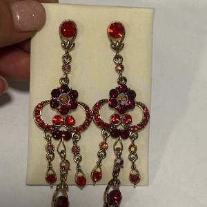Puede incluir: Un par de pendientes largos tipo candelabro en tono dorado con cristales rojos facetados dispuestos en patrones florales y circulares. Los pendientes incluyen elementos colgantes con piedras rojas pequeñas y un cristal rojo redondo en el poste.