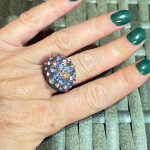 Puede incluir: Un anillo con una montura redondeada de tono oscuro, densamente incrustada con numerosos cristales pequeños, iridiscentes y multicolores en tonos azul claro, rosa y lavanda que reflejan la luz con un acabado brillante y destellante.