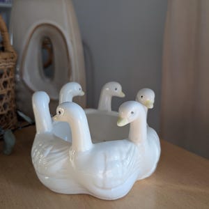 May include: A white ceramic bowl featuring a circular arrangement of four sculpted swan figures around the exterior. Each swan has a long neck, detailed wing feathers, and a small yellow beak, creating a continuous decorative rim around the basin.