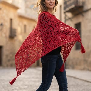 Puede incluir: Un chal triangular de ganchillo en color rojo brillante con un intrincado patrón calado y tres borlas rojas a juego en cada esquina. El chal se lleva sobre una camiseta negra y vaqueros azul oscuro, destacando su diseño texturizado.