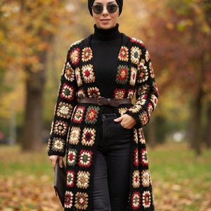 May include: A long, open-front cardigan featuring a multi-colored granny square crochet pattern in shades of red, green, cream, and black. The garment is worn over a black turtleneck sweater and black jeans, cinched with a brown leather belt.