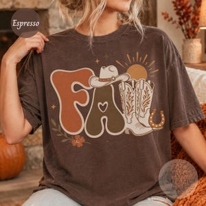 May include: A dark brown short-sleeve t-shirt featuring a graphic print with the word FALL in large, wavy letters. The design includes a cowboy hat, a pair of cowboy boots, a sun, a horseshoe, and floral accents in earthy tones.