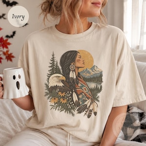 May include: An ivory t-shirt featuring a graphic print of a person with long dark hair, a bald eagle, pine trees, mountains, a sun, and flowers. The person wears a patterned shawl and feather earrings. The design uses earthy tones of brown, gold, and green.