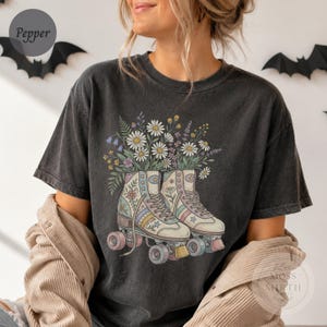 May include: A charcoal gray crew neck t-shirt featuring a graphic print of two vintage-style roller skates with pastel stripes and star accents. A bouquet of daisies, lavender, and green foliage emerges from the top of the skates.