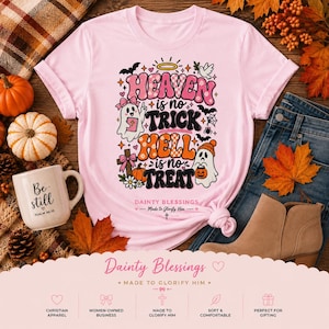 May include: A light pink t-shirt featuring a graphic with the text "HEAVEN is no TRICK HELL is no TREAT" and "DAINTY BLESSINGS Made to Glorify Him". The design includes two ghosts, a cross, a bat, a spider web, and a pumpkin in pink and orange.