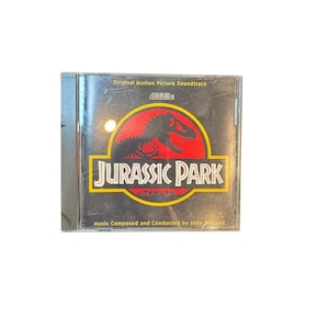 May include: A Jurassic Park original motion picture soundtrack CD in a clear plastic jewel case. The cover features a black background with a red circular logo containing a black dinosaur skeleton silhouette and the white text Jurassic Park.