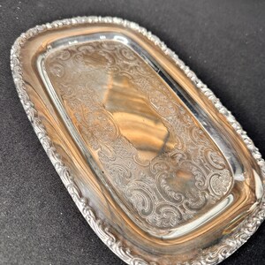 May include: An oval silver-toned metal tray featuring an intricate embossed scrollwork pattern along the border and a decorative etched design in the center. The tray has a polished reflective surface with a raised, scalloped edge detail.