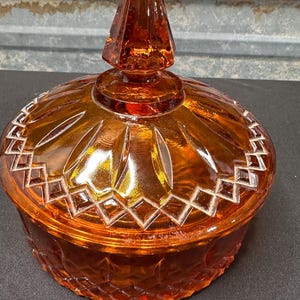 May include: A round amber glass candy dish with a matching lid. The lid features a pointed finial, a sunburst pattern, and a diamond-cut border. The base of the dish has a textured diamond pattern around the exterior, creating a reflective surface.
