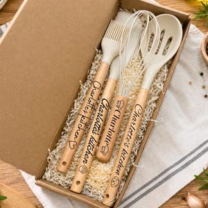 May include: A set of four kitchen utensils with cream-colored silicone heads and light wood handles, presented in a cardboard box with paper filler. Each handle is engraved with the text Charlotte's kitchen and various small icons.