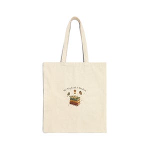 May include: A beige canvas tote bag featuring a central graphic of a stack of three books topped with flowers, a small lantern, and a butterfly. The text "My Weekend is Booked" is printed in a dark font above the illustration, surrounded by small stars.