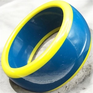 May include: A wide, circular bangle bracelet featuring a smooth, glossy finish. The exterior and interior surfaces are a deep, solid blue, while the top and bottom edges are accented with a vibrant, bright yellow trim that encircles the entire piece.