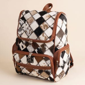 May include: A backpack featuring a patchwork design of black, white, and brown cowhide squares. The bag includes brown leather trim, multiple zippered compartments, and a small black rectangular patch on the front pocket with the letter H in silver.