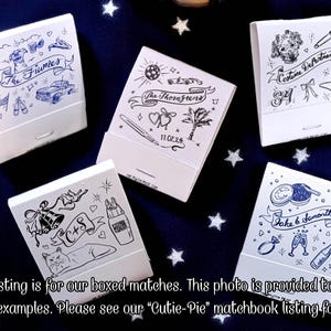 May include: A collection of white matchboxes with custom blue and black illustrations. Designs include names, dates, and various graphics like a car, dog, and champagne glasses. The matchboxes are displayed on a dark blue surface with white star accents.