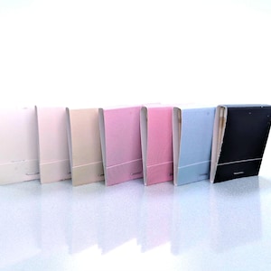 May include: A row of matchbooks in various colors, including white, pink, beige, and blue. Each matchbook has a rectangular shape with a textured surface and a contrasting bottom section. The matchbooks are arranged in a line against a white background.