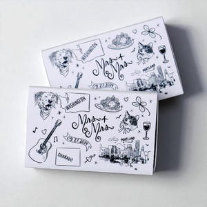 May include: Two white boxes with black line art illustrations. The boxes feature drawings of a dog, a cat, a guitar, and a cityscape. The text "Mrs. + Mrs." and the date "09.21.2024" are also present. The boxes are likely for a wedding.