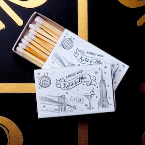 May include: Matchbox with a white and black illustration of a cityscape, a disco ball, and the text "A Perfect Match Katie & Tyler 11.1.24". The matchsticks have white tips and are inside the box.
