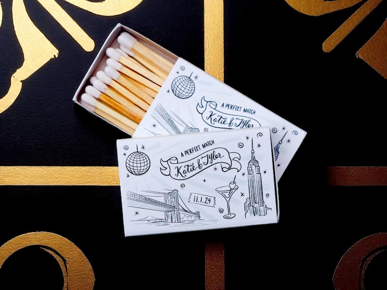 May include: Matchbox with a white and black illustration of a cityscape, a disco ball, and the text "A Perfect Match Katie & Tyler 11.1.24". The matchsticks have white tips and are inside the box.