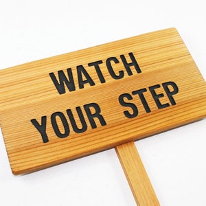 May include: A wooden sign with the text "WATCH YOUR STEP" in black lettering. The sign is attached to a wooden stick.