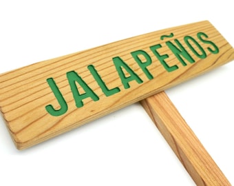 Puede incluir: Un letrero de madera con el texto "JALAPEÑOS" en letras verdes. El letrero está unido a una estaca de madera.