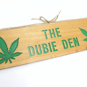 May include: A wooden sign with the text "The Dubie Den" in green lettering. The sign is decorated with two green marijuana leaf designs.
